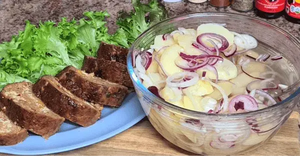 A vibrant potato salad layered with red and white onion slices, served next to golden-brown meatloaf and fresh greens.