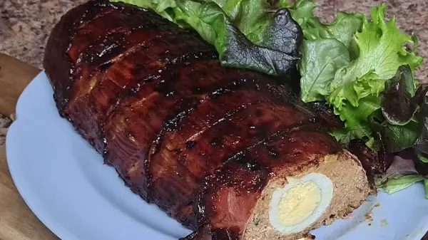 A golden-brown bacon-wrapped meatloaf sliced to reveal its cheesy, egg-filled center, garnished with crisp green lettuce.