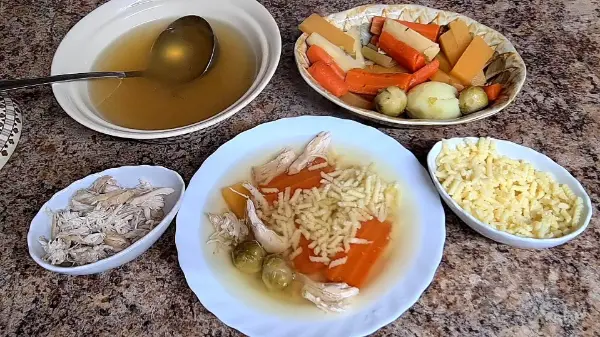 A comforting chicken soup served in a bowl with carrots, Brussels sprouts, shredded chicken, and tiny pasta, accompanied by side bowls of broth, vegetables, and pasta.