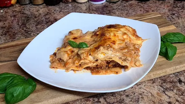 A slice of golden, cheesy lasagna served on a white plate with fresh basil leaves for garnish.