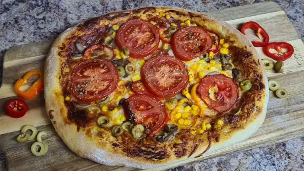A freshly baked pizza with a golden crust, topped with tomatoes, corn, olives, and cheese, served on a wooden board with sliced peppers on the side.