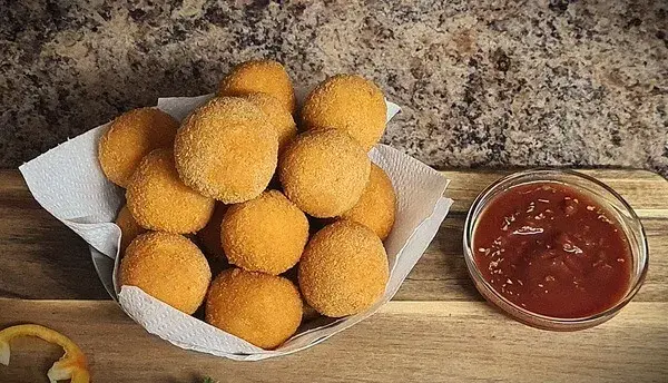 A wooden board featuring golden, crispy cheese balls stacked in a paper-lined basket, next to a small bowl of red dipping sauce.