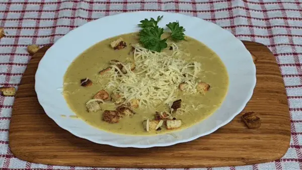 A creamy broccoli soup served in a white bowl, topped with grated cheese, crispy garlic croutons, and a sprig of fresh parsley. The bowl rests on a wooden board over a red-and-white checkered cloth, creating a warm and rustic presentation.