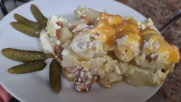 A comforting potato bake served on a white plate, featuring creamy layers of potatoes, melted cheese, ham, and sausage. Garnished with crunchy pickles on the side for a perfect flavor balance.