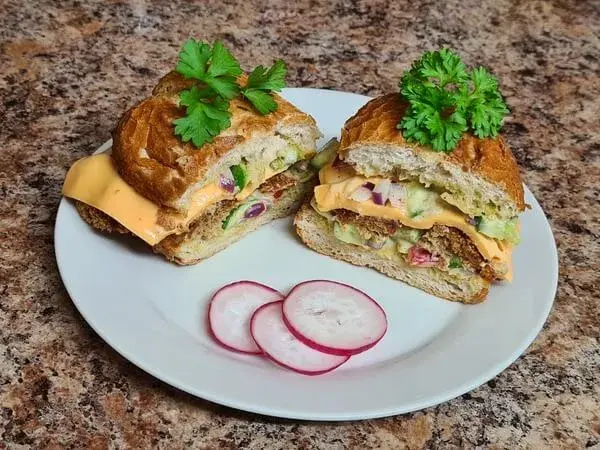 A crispy sandwich sliced in half, showcasing layers of golden meatball, melted cheese, fresh vegetables, and creamy mayo inside a toasted roll. Garnished with fresh parsley and served on a white plate alongside thinly sliced radishes.
