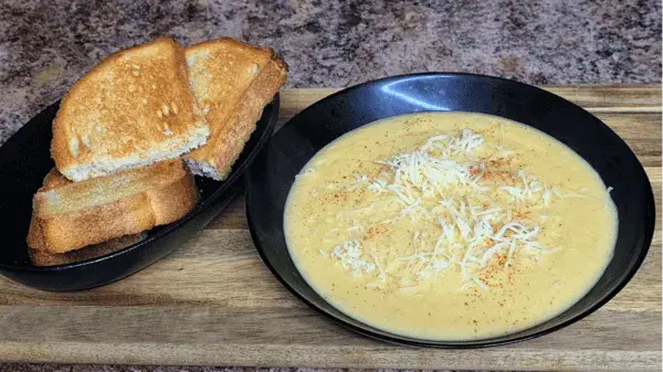 Rich creamy potato soup served in a black bowl, topped with shredded cheese, accompanied by golden toasted bread on a wooden tray.