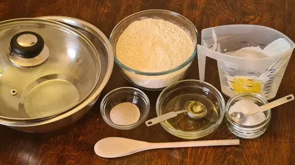 Ingredients for homemade bread including flour, yeast, water, oil, salt, bowls, and a wooden spoon arranged on a wooden table.