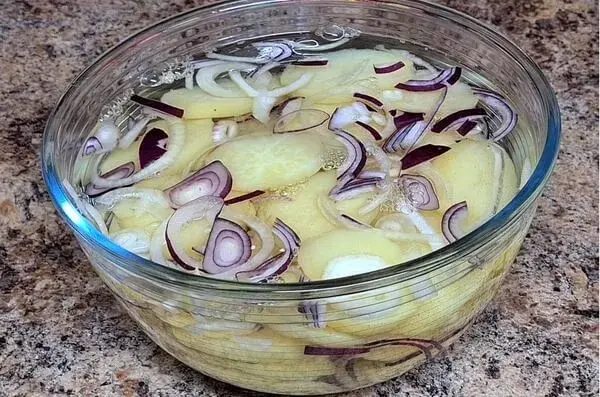 Glass bowl filled with sliced potatoes and red onions soaking in water on a kitchen counter.