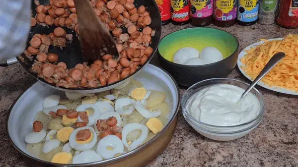 Layered potato casserole being prepared with boiled eggs, sausages, sour cream, and cheese.