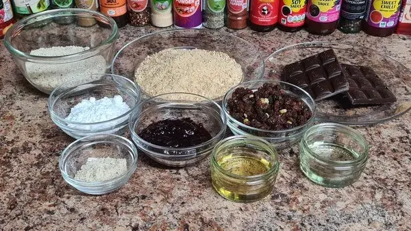 Ingredients for walnut balls: biscuit crumbs, ground walnuts, powdered sugar, dark chocolate, raisins, jam, vegetable oil, and spices neatly arranged in bowls on a kitchen counter.