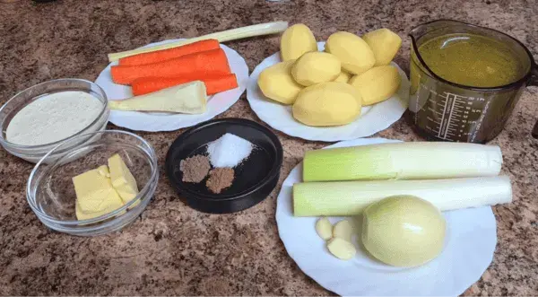 Ingredients for creamy potato soup: peeled potatoes, carrots, parsnip, leek, onion, garlic cloves, butter, cream, broth, and spices arranged neatly on a kitchen countertop.