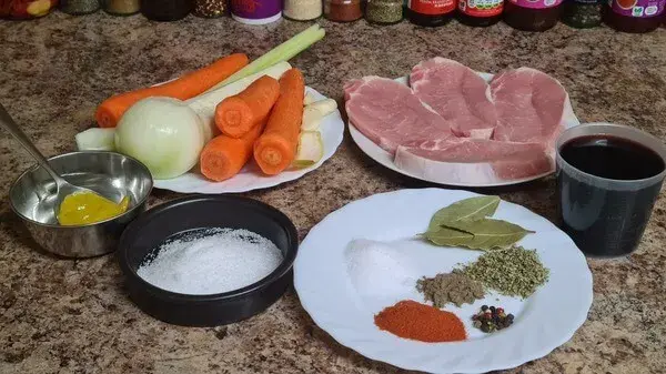 A selection of ingredients neatly arranged on a kitchen counter for a savory pork dish. A plate of raw pork medallions sits next to fresh vegetables, including carrots, onion, parsnip, and leek. A dish holds various spices like paprika, black pepper, bay leaves, and dried herbs, alongside a small bowl of mustard, a container of salt, and a cup of red wine.