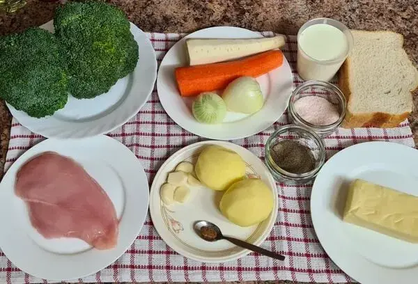 A variety of fresh ingredients laid out for a broccoli and chicken dish. The display includes raw chicken breast, fresh broccoli florets, potatoes, garlic, onion, carrot, parsnip, slices of bread, butter, milk, and small jars of salt and black pepper. Everything is arranged on a red-and-white checkered cloth.