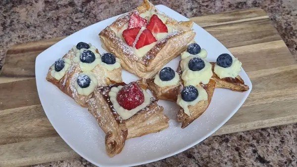 Golden puff pastry desserts decorated with vanilla custard and fresh strawberries and blueberries on a white plate.