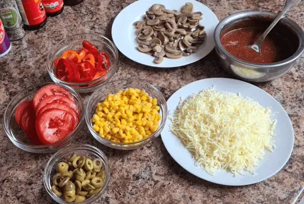 Ingredients for homemade pizza: sliced tomatoes, bell peppers, mushrooms, sweet corn, shredded cheese, and pizza sauce arranged neatly on a countertop.