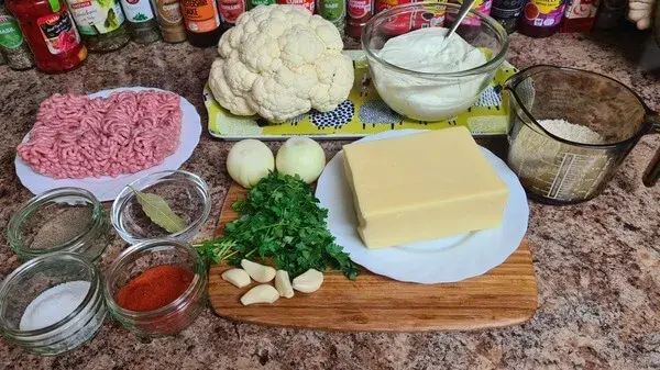 Ingredients for cheesy and creamy cauliflower casserole: fresh cauliflower, ground meat, large block of cheese, sour cream, onions, garlic, parsley, rice, paprika, bay leaf, salt, and spices arranged neatly on a kitchen countertop.