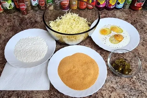 Ingredients for stuffed cheese balls with jalapeño: shredded cheese, sliced jalapeños, eggs, flour, and breadcrumbs arranged neatly on a kitchen countertop.