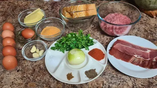 Ingredients for savory meatloaf with bacon, eggs, ground meat, onion, garlic, herbs, spices, breadcrumbs, cheese, and bread slices on countertop.