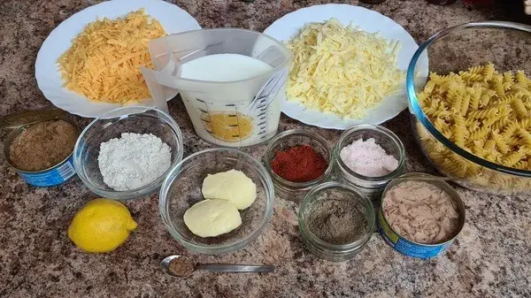 Ingredients for ultimate tuna mac & cheese: pasta, canned tuna, shredded cheese, milk, butter, flour, spices, lemon, and seasoning placed neatly on a kitchen counter.