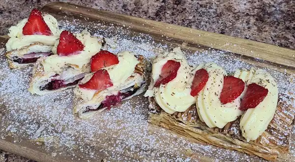 Close-up of a sliced cocoa roll with raspberry filling, vanilla pastry cream, and fresh strawberries on top, dusted with powdered sugar.