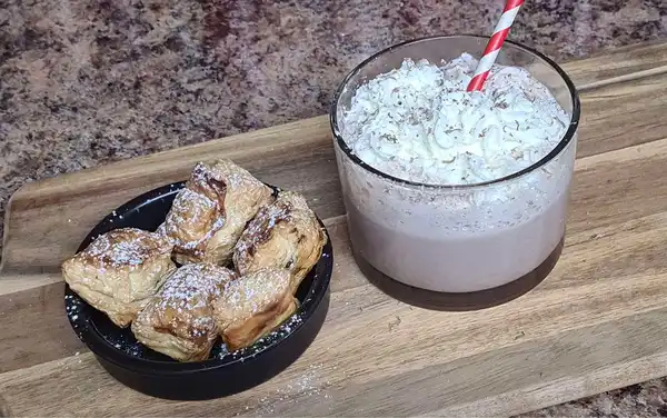 Mini puff pastry chocolate squares served in a black bowl, next to a glass of whipped cream-topped hot cocoa with a red striped straw.