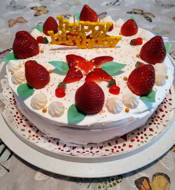 A beautifully decorated strawberry birthday cake with whipped cream frosting, halved strawberries, colorful sugar pearls, and a golden "Happy Birthday" topper.
