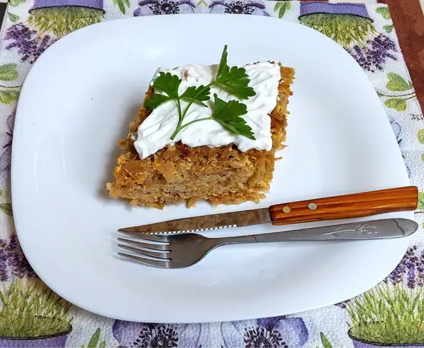 A hearty slice of stuffed sauerkraut casserole topped with sour cream and fresh parsley, served on a white plate with a rustic fork and knife on a lavender-themed napkin.