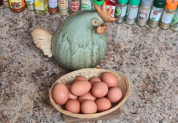 Quirky ceramic chicken watching over a basket of fresh brown eggs on a cozy kitchen counter, surrounded by spices.