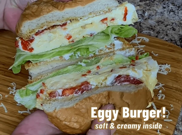 Close-up of a sliced Eggy Burger showing layers of creamy scrambled eggs, sweet red peppers, fresh lettuce, and mayonnaise inside a soft toasted bun.
