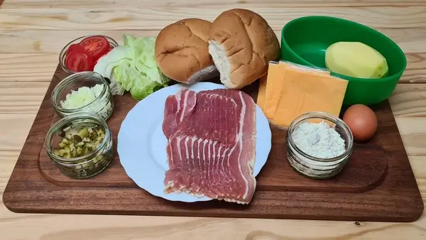 Ingredients for bacon burgers on a wooden tray, including lettuce, tomatoes, onions, pickles, burger buns, cheese slices, bacon, a peeled potato, egg, and flour.