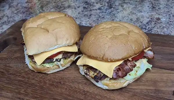 Two homemade bacon and cheese burgers with crispy potato patties, fresh lettuce, tomato, and a creamy sauce, served on a wooden board.