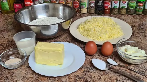 szöveg a hozzavalokhoz: Ingredients for cheese scone clouds: flour, grated cheese, butter, eggs, sour cream, milk, sugar, salt, and yeast arranged on a countertop.