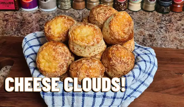 Golden, cheese-topped scone clouds on a rustic tray with herbs in the background – soft, savory, and freshly baked.