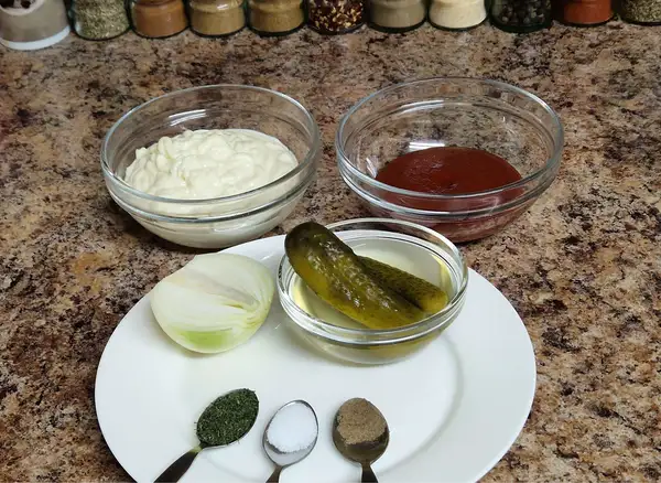 All the ingredients for homemade Thousand Island dressing arranged on a kitchen counter: bowls of mayonnaise and ketchup, a plate with sweet pickles in pickle juice, half an onion, dried dill, salt, and black pepper measured in teaspoons.