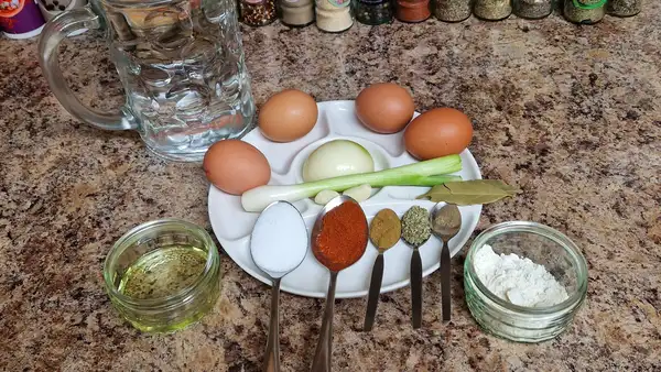 Ingredients for Hungarian egg drop soup arranged on a countertop: eggs, onion, scallion, bay leaf, spices, flour, oil, salt, and a glass of water.