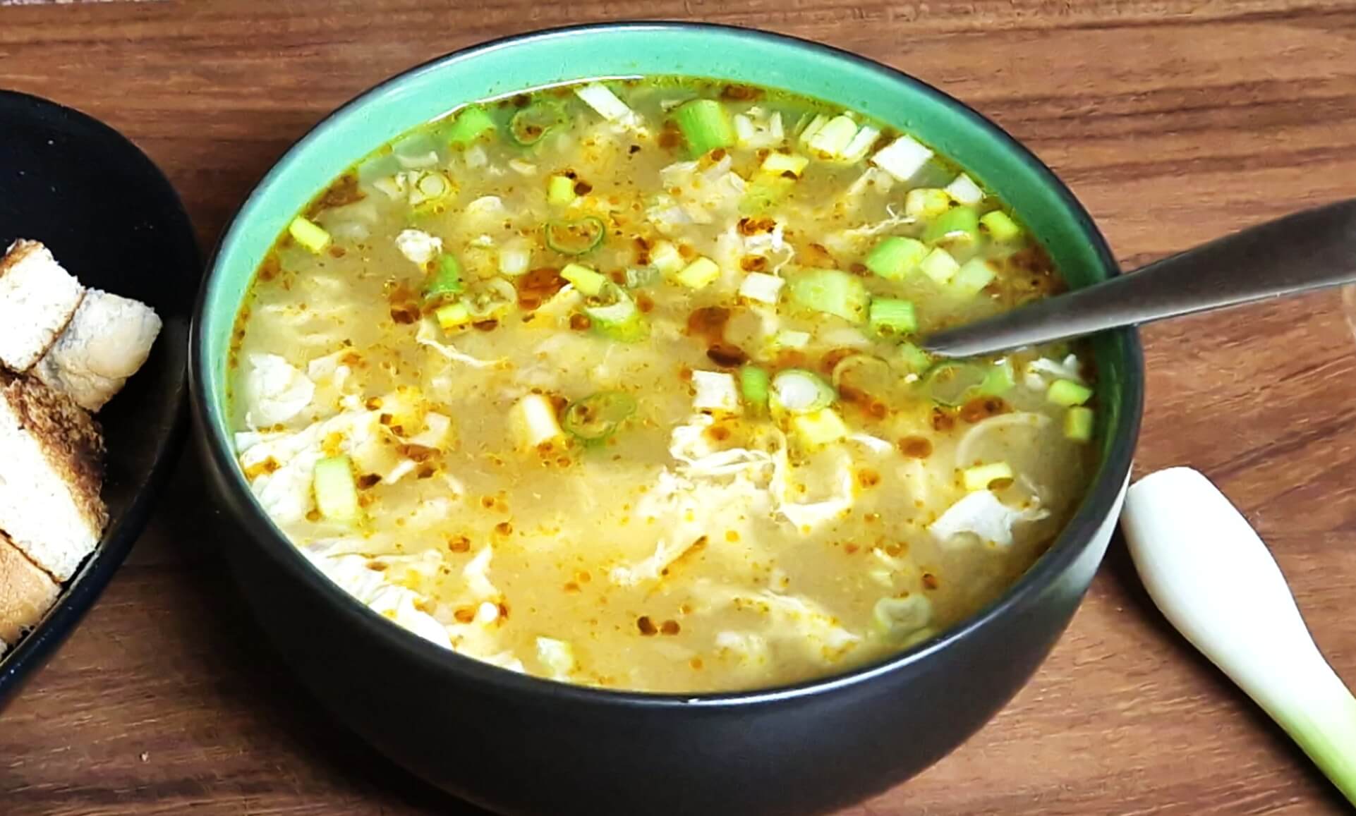 A bowl of Hungarian egg drop soup topped with sliced spring onions and chili oil, served with toasted bread cubes on a wooden board.