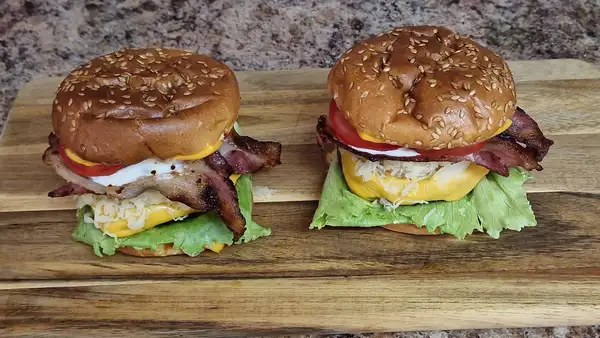 Close-up side view of a stacked BBQ pork kraut burger with melted cheese, fresh lettuce, tomato, sauerkraut, and seared ham inside a toasted bun.