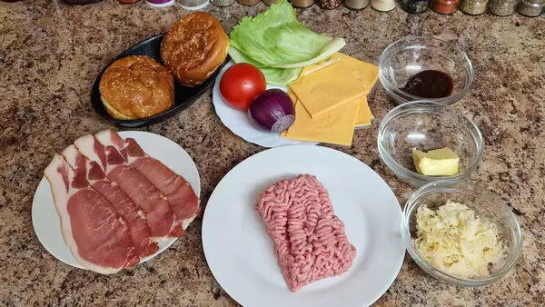 Ingredients laid out for BBQ pork kraut burgers, including ground pork, sliced tomato, onion, lettuce, cheese, ham, sauerkraut, mayonnaise, and sauces.
