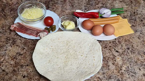 Ingredients laid out for a breakfast wrap: grated cheese, butter, bacon slices, tomato, pickled jalapeños, red sweet pepper, red onion, spring onion, smoked sausages, eggs, cheese slices, and a large tortilla on a kitchen counter.