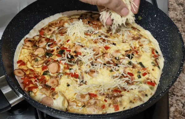 Grated cheese being sprinkled over a breakfast wrap filled with eggs, sausages, sweet red peppers, onions, and spring onions in a frying pan.