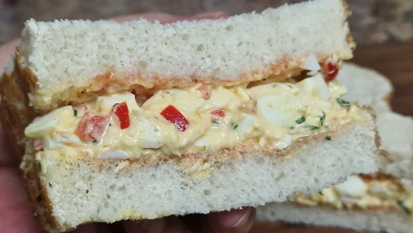 Close-up of a creamy egg salad sandwich filled with eggs, red bell pepper, herbs, and cheese between thick slices of white bread.