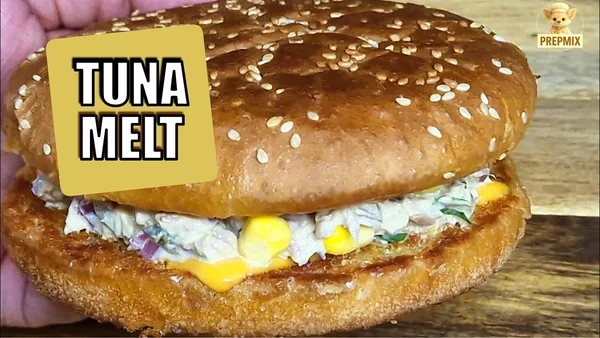 A golden, toasted sesame seed bun filled with creamy tuna, sweet corn, herbs, and melted cheese, served warm as a tuna melt sandwich.